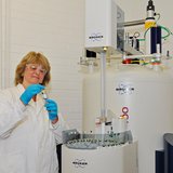 Eine Frau im weißen Chemie-Kittel steht vor einem Forschungsgroßgerät.
