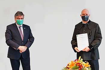 Zwei maskierte Personen stehen in einiger Entfernung, eine von ihnen hält eine Urkunde